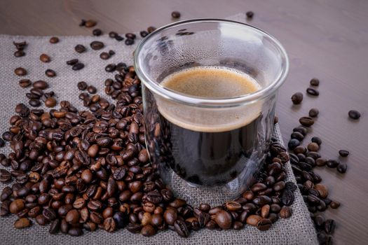 Close-up of glass cup with coffee and pile of roasted coffee beans on piece of burlap on wooden surface. Heat-resistant coffee glass with double walls on table. Selective focus.