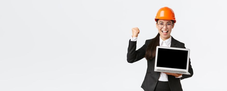 Successful winning businesswoman, construction manager or engineer at factory showing profit chart, diagram at laptop screen, fist pump in rejoice, standing pleased over white background.