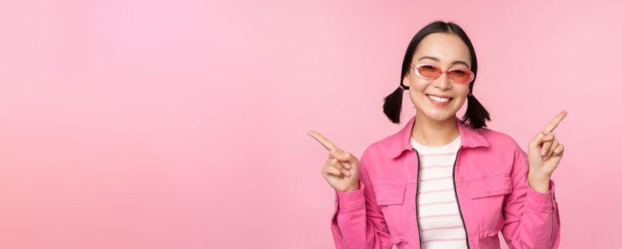 Choice. Stylish korean girl, asian female model points fingers sideways, shows two variants, product advertisement, demonstrating items, standing over pink background.