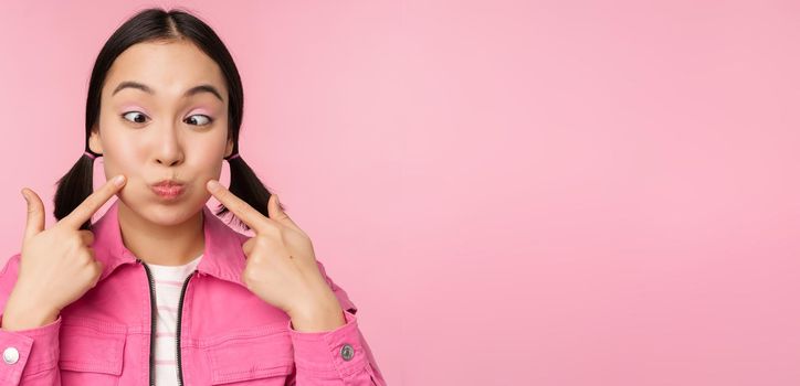 Close up portrait of young asian girl showing her dimples, poking cheeks silly and making funny faces, standing over pink background.