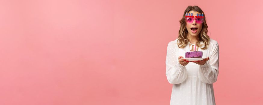 Holidays, spring and party concept. Excited, happy attractive blond woman in white dress, wearing funny b-day glasses, holding birthday cake with lit candle, making wish smiling camera.