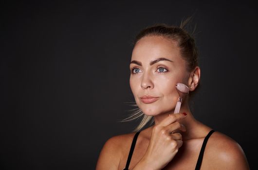 Close-up of a beautiful middle aged Caucasian woman massaging her face using a jade stone roller massager, isolated over black background with copy ad space