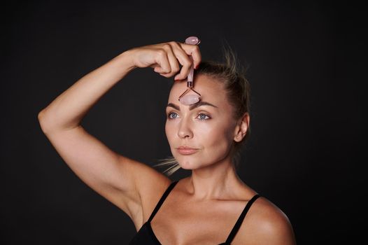 Face portrait of a pretty Caucasian woman wearing black underwear doing a lymphatic drainage facial massage with a roller massager, isolated over dark background. Beauty spa treatment, copy space