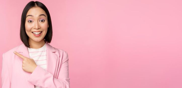 Portrait of asian business woman, saleswoman in suit pointing finger left, showing banner advertisement, smiling and looking professional, pink background.