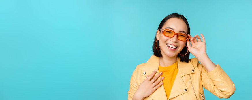 Close up portrait of stylish asian woman in sunglasses, laughing and smiling, looking happy, posing in trendy clothes over blue background.
