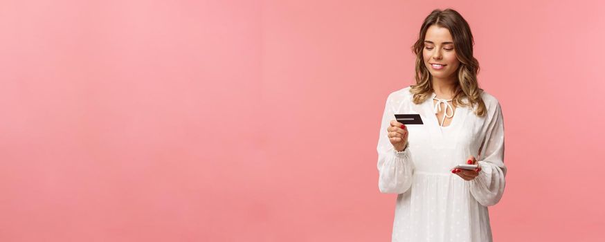 Portrait of attractive and cute blond girl with short curly hairstyle, white dress, holding smartphone and credit card, insert digit numbers to register in shopping app, buy online, pink background.