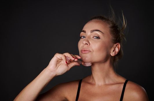 Anti-aging, smoothing, rejuvenation beauty therapy. Middle aged beautiful woman with healthy fresh clean skin massaging her face with jade stone roller massager. Studio shot