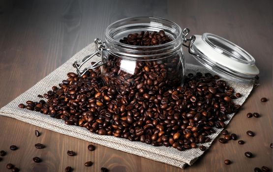 Close-up of glass jar with roasted coffee beans on pile of beans on wooden surface. Selective focus.
