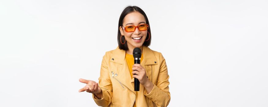 Portrait of beautiful asian woman in sunglasses, stylish girl singing, giving speech with microphone, holding mic and smiling, standing over white background. Copy space