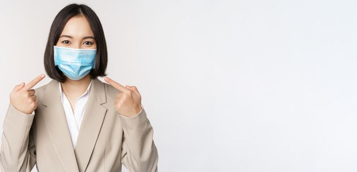 Coronavirus and business people concept. Asian female entrepreneur pointing fingers at medical face mask at workplace, standing over white background.