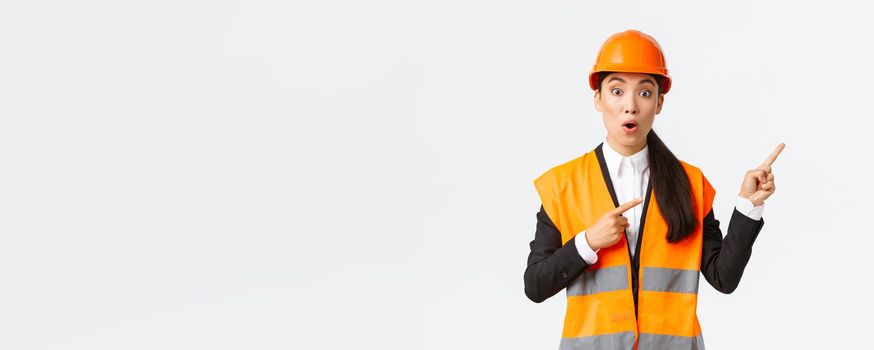 Building, construction and industrial concept. Impressed and surprised asian female engineer asking question about interesting product or object. Architect pointing fingers upper right corner.