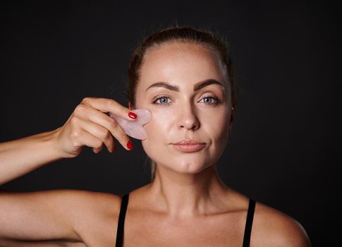 Studio shot of a pretty Caucasian woman doing a lymphatic drainage facial massage with a jade stone gua-sha massager. Beauty spa treatment. Close-up. Anti-aging, smoothing, rejuvenation beauty therapy