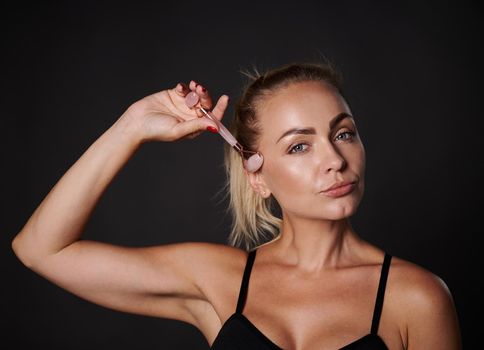 Studio shot of a pretty woman doing a lymphatic drainage facial massage with a jade stone roller massager. Beauty spa treatment. Close-up. Anti-aging, smoothing, rejuvenation beauty therapy