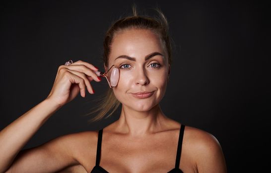 Close up of middle aged European woman with healthy glowing tanned skin, massaging face with jade roller isolated over black background with copy ad space