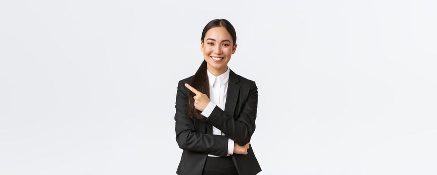 Happy professional asian female manager, businesswoman in suit showing announcement, smiling and pointing finger left at product or project banner, standing white background.