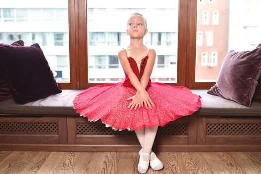 Smiling baby girl dreaming to become professional ballet dancer, classical dance school. the little ballerina does stretching sitting against the window