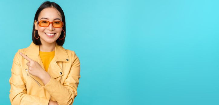 Stylish smiling asian woman in sunglasses, pointing finger left, showing advertisement banner, standing in trendy yellow coat over blue background.