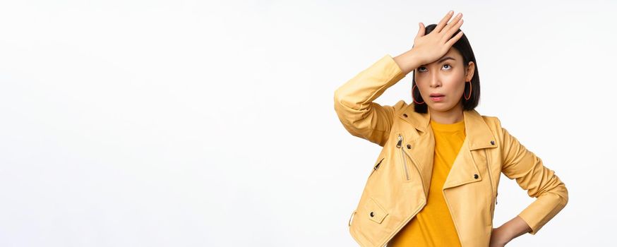 Annoyed and tired asian girl, slap forehead, facepalm and eyeroll with bothered face expression, standing in casual clothes over white studio background.