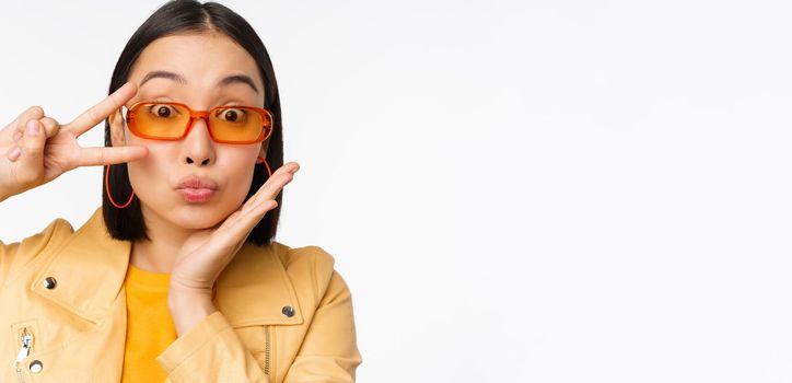 Close up portrait of young asian woman, stylish girl in sunglasses, showing peace, v-sign and smiling, standing over white studio background.