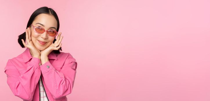 Portrait of stylish cute asian girl, smiling and touching face, looking up dreamy, thoughtful look, standing over pink background.