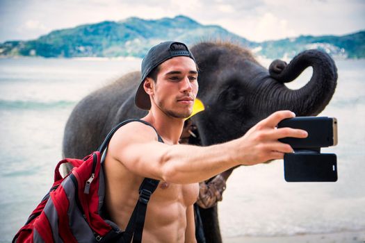 Confident young muscular Hispanic male traveler with naked torso on cap and backpack, taking selfie with elephant on mobile phone, standing on ocean beach during trip in tropical country