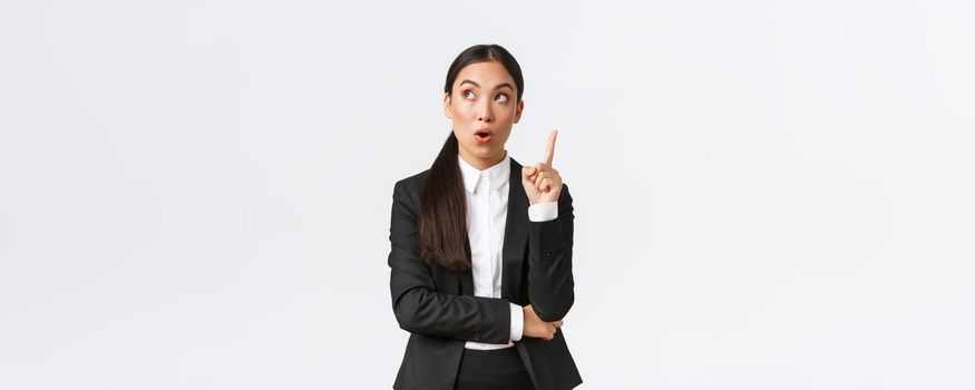 Smart and creative asian businesswoman have excellent idea, raising finger up and looking upper left corner thoughtful, suggest solution, make plan, saying thought over white background.