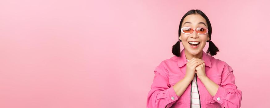 Portrait of excited japanese girl in sunglasses, celebrating, achieve goal, gasping amazed and smiling, standing over pink background.