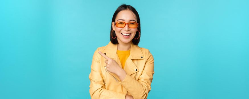 Smiling asian woman in sunglasses, pointing finger left, showing banner, logo or advertisement, standing over blue background. Copy space