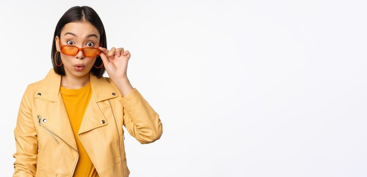 Portrait of asian brunette woman in stylish sunglasses, looks surprised and impressed at camera, checking out big news, wow face expression, standing over white background.