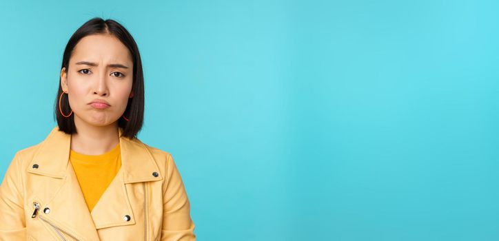 Close up of disappointed asian girl sulking, grimacing and frowning displeased, standing moody against blue background. Copy space