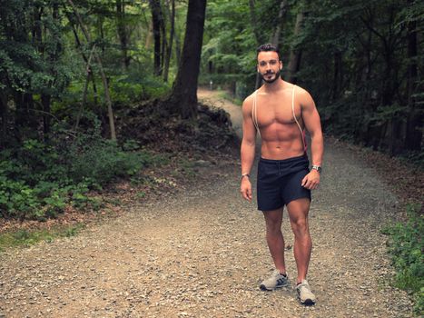 Handsome young man shirtless outdoors hiking on park path with backpack on shoulder. Full length shot