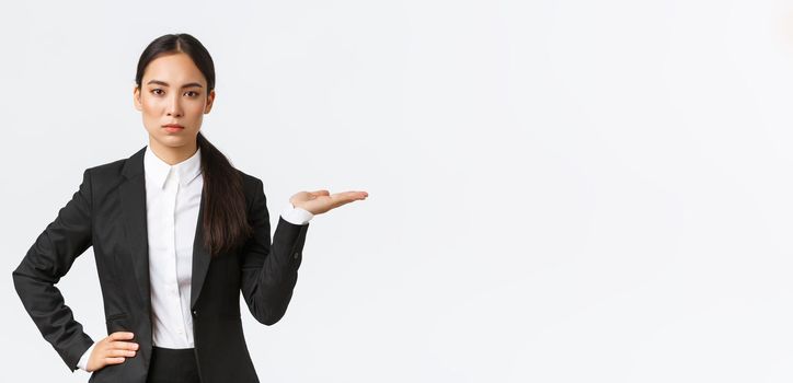 Serious-looking determined young asian businesswoman introduce her project, showing something on palm, holding hand right on blank space, as introduce product, standing in black suit white background.