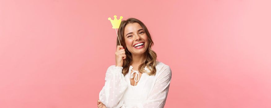 Spring, happiness and celebration concept. Close-up portrait of charming smiling, lovely blond girl holding small queen crown on stick, laughing joyfully, feel empowered and happy, pink background.