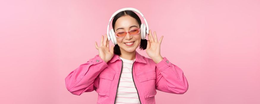 Dancing stylish asian girl listening music in headphones, posing against pink background.