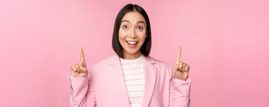Enthusiastic corporate worker, asian business woman pointing fingers up and smiling, showing advertisement, logo, standing over pink background.