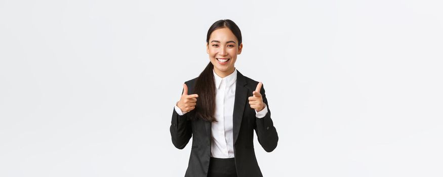 Confident smiling asian saleswoman in black suit showing thumbs-up, guarantee quality of product or providing best service. Sales manager trying sell something to client, white background.