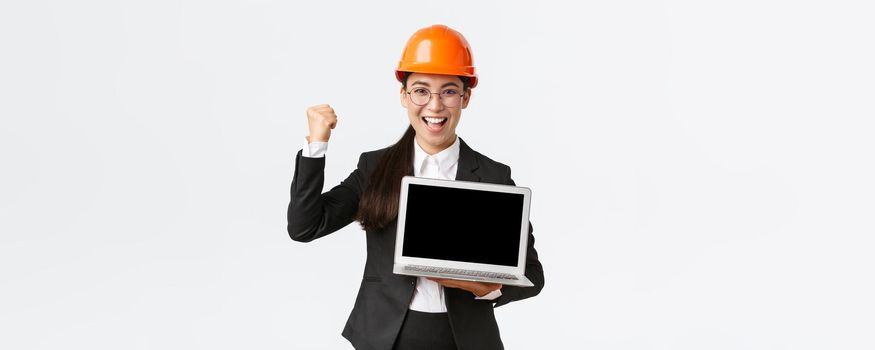 Successful winning businesswoman, construction manager or engineer at factory showing profit chart, diagram at laptop screen, fist pump in rejoice, standing pleased over white background.