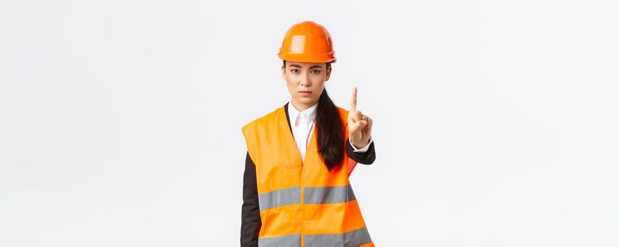 Serious-looking confident and displeased asian female engineer shaking finger, scolding construction worker for acting dangerous, prohibit and forbid action at building zone, white background.
