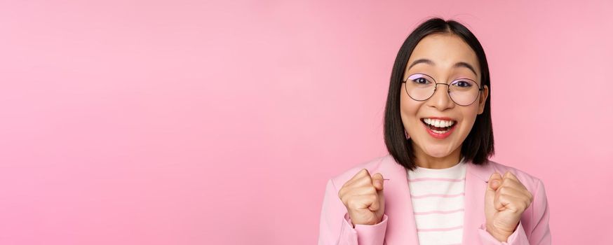 Close up portrait of asian businesswoman cheering, rooting for, looking with hope and excitement at camera, smiling and laughing, standing over pink background.