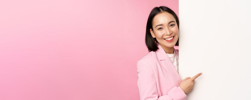 Image of asian female entrepreneur in suit, pointing finger at board, showing smth on white wall, demonstrating chart or information, standing over pink studio background.