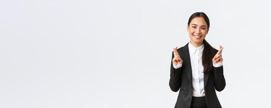 Hopeful businesswoman awaiting for promising results, cross fingers good luck and smiling optimistic. Happy saleswoman in black suit making wish, anticipating good news, white background.