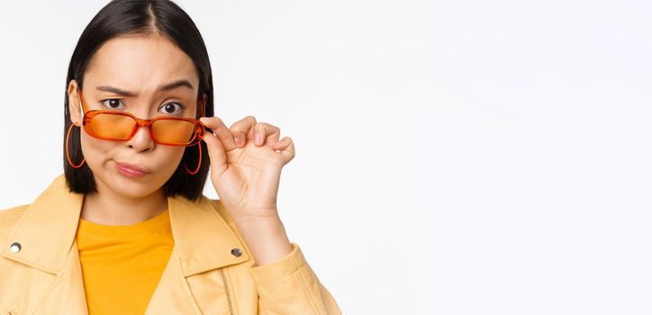 Image of young asian stylish woman, looking from under sunglasses with skeptical, disbelief face, express doubt, standing over white background. Copy space