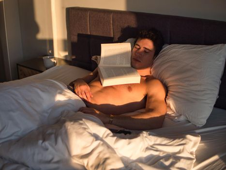 Young man lying in bed and reading a book then falling asleep