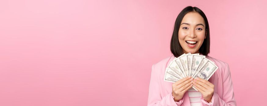 Finance, microcredit and people concept. Happy smiling asian businesswoman showing dollars money, standing in suit against pink background.