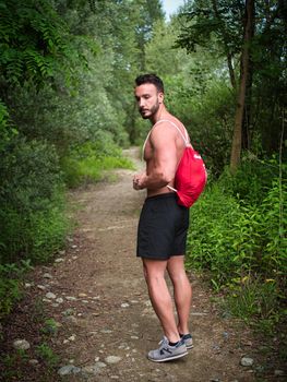 Handsome young man shirtless outdoors hiking on park path with backpack on shoulder. Full length shot