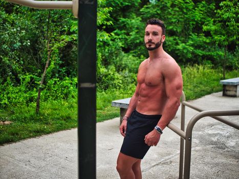 Attractive shirtless young man exercising and working out in outdoor gym in city park, leaning on equipment