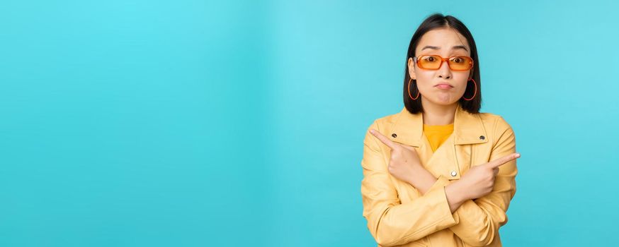 Indecisive asian woman in sunglasses, makes choice between two variants, points sideways, looks puzzled, stands over blue background.