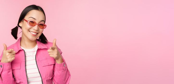 Close up portrait of modern asian girl in sunglasses smiling, pointing fingers at camera, praise you, inviting or complimenting, standing over pink background.