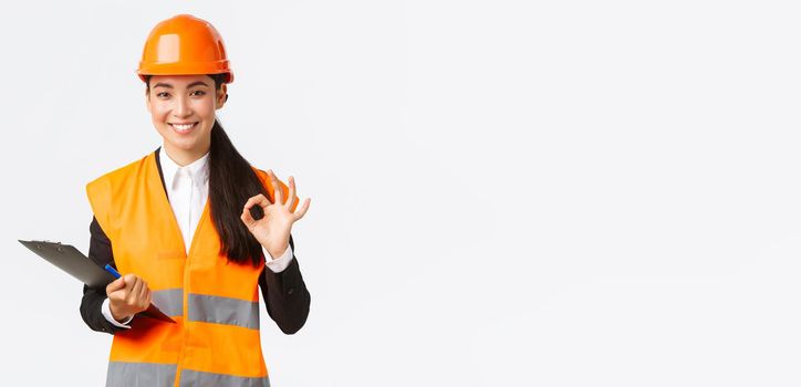 Satisfied smiling asian female architect, construction engineer in helmet and reflective jacket, showing okay gesture, approve and give permission for builing works, finish inspection.