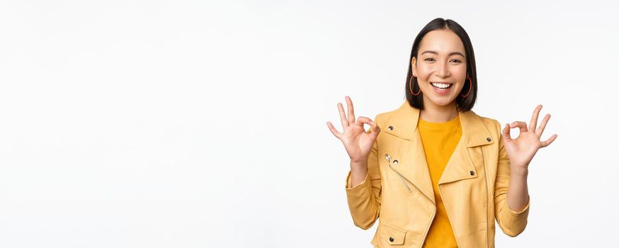 Happy beautiful asian girl shows okay, ok recommending sign, pleased by smth, praising quality, standing over white background. Copy space